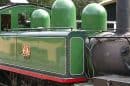 Vintage green and black steam train at a station with red and yellow carriages nearby.