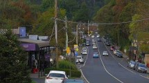 Main street in a mountain village with parked cars and shops, surrounded by forested hills.